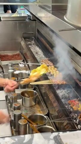 The video shows a person grilling meat skewers over hot coals. The person is wearing a hair net and is using metal skewers to hold the meat. There are several skewers of meat on the grill, some of which are already cooked and browning. There are also containers of spices and sauces nearby, as well as a metal tray with more meat. The environment appears to be a professional kitchen or food stall. The grilling is taking place on a long, narrow grill with a metal grate over a bed of hot coals. Smoke is rising from the grill as the meat cooks. The person is actively turning and adjusting the skewers to ensure even cooking.