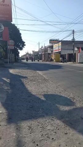 A straight, paved road stretches into the distance under a clear sky. On the left side of the road, a tall red sign displays text in white, and below it, a smaller red sign shows a cartoonish image of a smiling cow with the word "BAKSO" written on it. A mature tree stands behind these signs, its leaves casting a dappled shadow on the road. The right side of the road features several buildings, one with a yellow awning and another with a white facade. A few motorcycles are visible on the road, some with riders, moving away from the viewer. Shadows of trees and possibly people are cast across the rough, gravelly shoulder of the road in the foreground. Overhead, a tangle of electrical wires crisscrosses the sky. The overall impression is that of a sunlit day in a rural or suburban area with moderate traffic.