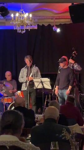 The video shows a jazz band performing on stage in front of an audience. The band consists of a drummer, a flute player, a saxophone player, a guitarist, and a bassist. The drummer is seated behind an orange drum set, wearing glasses and a gray shirt. The flute player stands at the center, holding a flute, dressed in a patterned shirt and dark pants. The saxophone player stands to the right, wearing a black sweatshirt, jeans, and a baseball cap. The guitarist is seen to the left, playing a red electric guitar while wearing glasses. The bassist is partially visible behind the saxophone player, standing with his bass. The stage is decorated with a chandelier, blue and red lighting, and black curtains. The audience sits at tables, watching the performance. The room appears to be a restaurant or club.