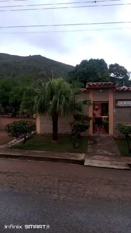 The video shows a man standing in the doorway of a light brown and tan building. The building has a sign that reads "NO ME OLVIDES". The camera pans to the right, revealing a house adjacent to the first building, covered in pink flowers. There are trees, bushes, and a mountain in the background. The sky is overcast and the street is wet. The bottom of the screen reads "Infinix SMART 8".