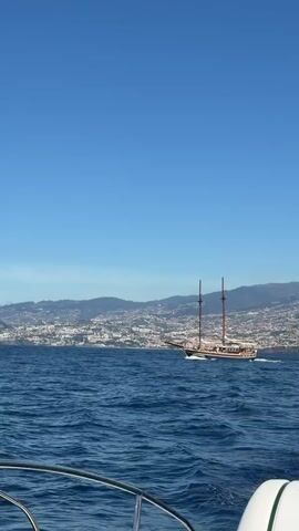 The video shows a boat moving on the ocean on a clear, sunny day. The camera is positioned on a boat, with the railing and part of the boat's side visible in the foreground. In the middle ground, another larger boat with two masts is traveling in the same direction. In the background, there is a coastline with mountains and what appears to be a city. The sky is a clear blue, and the water is a deep blue with whitecaps.