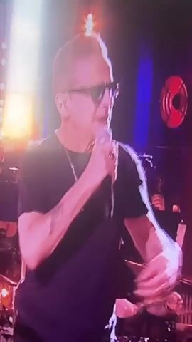 The video shows a man wearing sunglasses and a black t-shirt, holding a microphone and singing. He has short, light-colored hair and appears to be on a stage with other people in the background. There are lights and what appear to be musical instruments. The lighting is dim and colorful, with pink and purple hues dominating.