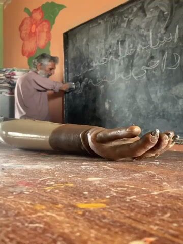 An older man with gray hair and beard wearing a light brown shirt stands in front of a chalkboard with Arabic script, smiling and spreading his arms. On a wooden table in the foreground is a prosthetic arm with a dark brown hand and metal finger tips. The room has a peach-colored wall decorated with a painting of an orange flower. There are papers stacked to the left of the chalkboard.