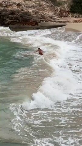 The video shows a person standing in the ocean near a beach on a bright, overcast day. The person is wearing red swim trunks and is waist-deep in the water. Small waves are breaking around them, creating white foam on the surface. Behind the beach, there is a rocky cliff covered in vegetation. The water is a mix of turquoise and tan colors, with the tan appearing closer to the shoreline. The waves are small but visible. The camera appears to be positioned at a high angle, possibly from an elevated viewpoint along the coast.