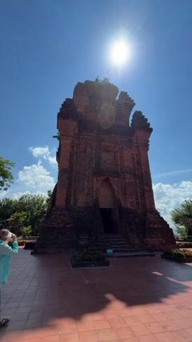 A person wearing a light blue top and dark pants stands on a red tiled plaza, holding a phone and taking a photo of a tall, ancient brick structure. The structure has a dark, arched entrance at the base and appears weathered and somewhat dilapidated, with vegetation growing at its top. The sun is high in the bright blue sky, creating a strong glare and casting a shadow of the structure on the plaza. Trees are visible to the left and right of the structure. The scene conveys a sense of historical exploration and tourism on a sunny day.
