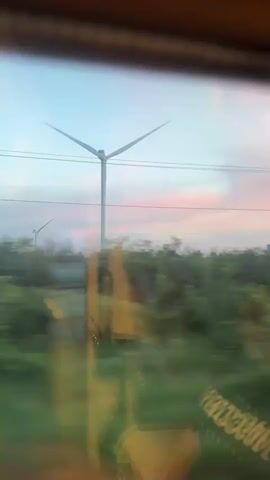 The video shows a view from a moving vehicle, likely a train, looking out at a landscape. The primary subject is a tall, white wind turbine with three blades, which are blurred due to motion and appear to be slowly rotating. In the background, another, smaller wind turbine is partially visible.

The scene is set in a rural or semi-rural area with lush green fields and trees. Power lines stretch across the frame, intersecting with the sky. The sky itself is a prominent feature, displaying a soft gradient of colors, with a pale blue at the top fading into hues of pink and white, suggesting either dawn or dusk. The overall mood conveyed is tranquil and serene, with the motion of the vehicle adding a dynamic element to the otherwise still landscape.

There is no visible text in the media. The time of day is indicated by the soft, pastel colors of the sky, typical of sunrise or sunset. The location is identified as Verkhnegoveinyi, Russia, which implies a northern latitude where such landscapes and renewable energy installations might be found. The focus on the wind turbine highlights a theme of sustainable energy.