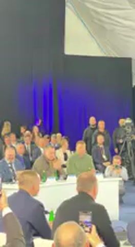 The video shows an indoor gathering of numerous people, mostly men, in what appears to be a large tent or event hall. Many attendees are seated at white tables, some with bottles and other items, while others stand in the background. A blue backdrop is visible behind a stage area, with bright white fabric forming the ceiling.

At the beginning of the video, the audience is seen, and a man in a black long-sleeved shirt (likely Volodymyr Zelenskyy, given the context provided) walks forward from the left side of the frame towards the center. He approaches another man dressed in a blue suit who is standing on a small elevated platform or stage. The man in the blue suit holds a red, rectangular object, possibly a book, plaque, or award. As the man in black approaches, the man in the blue suit extends the object towards him.

The man in black accepts the item, and the two men stand together, facing the audience. There is visible applause from the crowd, and a camera on a tripod is positioned to record the event. Other individuals, possibly security or staff, are seen standing behind the main figures. The atmosphere appears to be one of recognition or presentation during an official event. No discernible text is visible on banners, screens, or other objects within the frame. The location context provided is Barvinok, Ukraine.