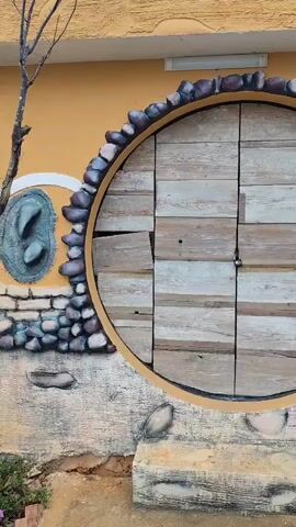 The video displays a static shot of an exterior wall, prominently featuring a large, circular wooden door. The door, constructed from light-toned horizontal wooden planks, is framed by a golden-brown ring and secured by a small, dark padlock or latch in its center.

The wall surrounding the door is elaborately decorated with natural elements. A border of rounded, dark gray stones encircles the door's frame, extending downwards and to the left, where they are integrated into a textured, light-colored stucco-like base. To the immediate left of the circular door, an irregular, raised section of the wall also incorporates embedded stones and three smooth, dark gray, oval-shaped features. The upper part of the wall is painted a warm, textured yellow or ochre.

In front of the door, a low, light-colored concrete step or platform is visible, with the ground appearing as dirt beyond it. A thin, bare tree branch can be seen on the far left of the frame. To the right of the circular door, on the yellow wall, a colorful mural is partially visible. It includes blue text that appears to read "Đồi M..." or "Đôi M..." and elements resembling foliage or a tree.

The scene is captured in natural daylight, indicating clear weather conditions. The overall aesthetic suggests a whimsical, fantasy-inspired architectural design, likely part of a themed attraction, resort, or unique dwelling in Lạc Dương, Vietnam. No people or activity are present in the shot.