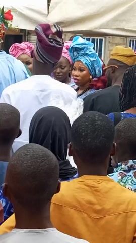 Here is a detailed description of the video:

**Scene:** The video captures a gathering of people, likely at a social event or celebration.

**People:** The focus is on a group of individuals, possibly attendees of the event. The women are prominently dressed in traditional attire, with elaborate headwraps in varying colors, including blue, pink, and striped patterns. A man is holding a microphone and seems to be addressing the group. Others are observing or listening. Children are also part of the crowd, visible in the foreground with their backs to the camera.

**Objects:** 
- Microphones: The man's is the only microphone that appears in the shot.
- Headwraps: Various colorful and intricately designed headwraps adorn the women's heads.
- Clothing: The attire ranges from traditional dresses and gowns to more casual shirts.
- Accessories: A necklace and sunglasses.

**Actions:** The main action is the man speaking into the microphone, possibly making an announcement or giving a speech. People are listening, observing, and interacting with each other.

**Interactions:** There is a sense of community and engagement among the people present. They are gathered closely, suggesting a shared interest or involvement in the event.

**Environment:** The event appears to be held outdoors or in a semi-outdoor space, as evidenced by the presence of natural light and the structure visible in the background. The setting seems to be a residential or public area.