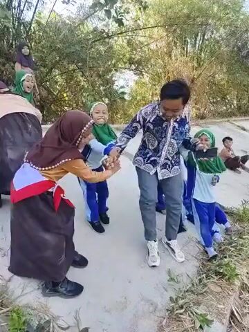 A group of children, some wearing hijab, and an adult man are outdoors. The man is wearing a patterned shirt and gray pants. The children are dressed in school uniforms, some with green hijabs. They appear to be engaged in some form of activity or game, possibly a dance or a group exercise, as they are holding hands and moving. The background shows lush vegetation, including bamboo trees, and a dirt or sandy path. The lighting suggests it is daytime.