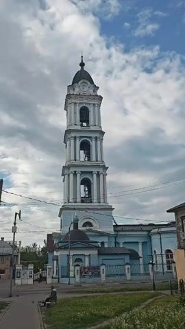 The video shows a blue and white church with a tall bell tower against a partially cloudy sky. The tower is multi-tiered and has a clock face near the top. A person is sitting on a bench near the church, and a car drives by on the street. Other buildings are visible in the background, including a two-story wooden structure with an exposed window frame. There are lampposts and power lines along the street. Grass grows on the lawn between the street and the church. The church is surrounded by a fence. The bells are ringing.