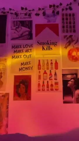 The video depicts the interior of a room with walls covered in a variety of posters, prints, and art pieces. The lighting in the room is predominantly purple, possibly from LED or decorative lights. The camera moves from left to right, showing different sections of the walls and their decorations. One wall has a collection of framed paintings. At one point, a white fan is visible in the room. The scene also contains a bed with a dark-colored blanket. The room has a casual, personalized aesthetic. Music plays in the background.