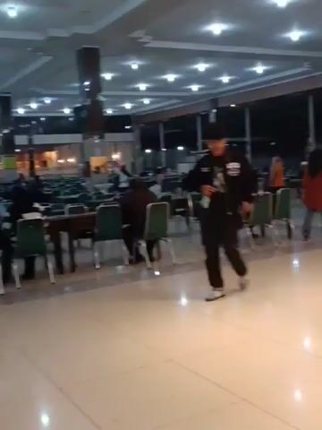 The video shows the interior of a brightly lit establishment, possibly a restaurant or food court. In the foreground, a stainless steel counter is visible, stacked with plates. To the left of the counter, a person in an orange shirt is interacting with a food display. Further left, a person in a black shirt and cap walks past. In the background, several other individuals are present, some seated at tables, and others appear to be working. The environment is clean and well-maintained, with a modern aesthetic characterized by bright lighting and polished floors. The overall impression is of a busy dining area during evening hours.