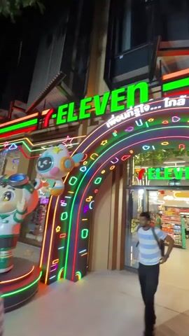 A person walks towards a brightly lit 7-Eleven store entrance. The entrance is decorated with a colorful neon archway featuring abstract shapes and cartoon characters. The store's interior is visible through the glass doors, showcasing shelves stocked with various products and other shoppers. The store appears to be open 24 hours, as indicated by signage on the doors.