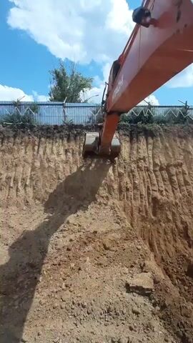 A Hitachi excavator digs into a dirt embankment under a bright blue sky with scattered clouds. The excavator's orange arm and bucket are prominent in the foreground, casting a shadow on the excavated earth. The embankment has distinct horizontal layers from previous digging. A metal fence with diamond-shaped openings runs along the top of the embankment, with some green foliage visible behind it. The scene appears to be a construction or landscaping site.
