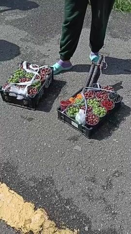 The video shows an outdoor market scene where two crates filled with various types of grapes are displayed on the asphalt. The crates contain grapes of different colors, including green, red, and dark purple. A person's legs are visible in the foreground, wearing green sweatpants and light blue and purple socks. There are other people in the background, also appearing to be at the market. The ground is asphalt with a yellow painted line. The overall impression is of a vendor selling fruit at an outdoor stall.
