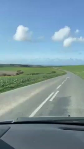 A vehicle is driving on a paved road through a rural landscape. The sky is blue with scattered white clouds. The road curves through green fields. Some areas of the fields are brown. In the distance, a line of trees is visible. The camera is inside the vehicle, looking out the windshield.