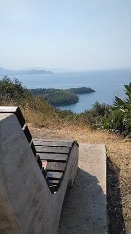 The video shows a view from a bench overlooking a bay. The bench is made of concrete and wood. The bay is calm and blue, with a few boats visible. Lush green trees line the shore. The sky is clear and blue. The overall scene is peaceful and scenic.
