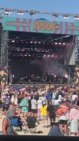 The video shows a festival stage from a crowd's perspective. A band is performing on stage, with members visible behind smoke. The stage is set up outdoors, with a large screen displaying images and the festival's name, "МУЗЫКАЛЬНЫЙ ПЛЯЖ 26 ФЕСТИВАЛЬ." The crowd is a mix of adults of various ages and ethnicities, with many people sitting in chairs or standing. Some are holding umbrellas. The ground appears to be sand. The sky is blue and clear.