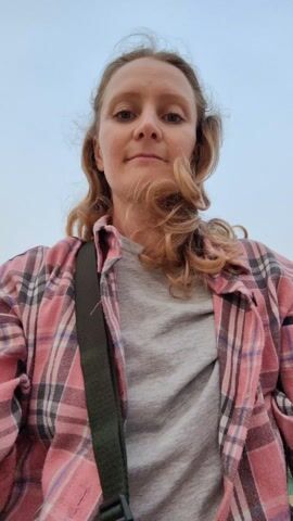 A woman in a pink plaid shirt is lying on the sand and smiling up at the camera. A green trash can is visible in the background. Someone wearing blue shorts walks past the camera. The sky is an overcast blue color.