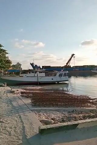 The video shows a harbor with several boats. In the foreground, there is sand and what appears to be construction materials, possibly rebar. A white boat is docked on the left side of the frame, partially on the sand. In the background, there are larger vessels. One is a cargo ship carrying containers, with a crane extending upwards. Another white passenger boat is visible near the cargo ship. The water is calm, reflecting the sky, which is blue with scattered clouds and illuminated by the sun, suggesting it is daytime. The overall scene depicts a working harbor with ongoing activity or preparation.