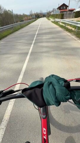 The video shows a first-person perspective of a person riding a red bicycle down a paved road with a white line in the middle. The road is bordered by a metal guardrail on either side. A sign in Serbian, indicating a village or town called Džiminica, is visible on the right side of the road. The view is mainly of the road ahead, with some glimpses of the surrounding landscape, which includes trees and houses. A person on a black bicycle then comes into view from the left, riding a wheelie. The person is wearing all black, including a t-shirt, pants, and shoes. The person riding a wheelie passes the camera on the left before moving out of the scene. The first-person view of the bicycle moving along the road resumes after the wheelie rider has passed. The sky is overcast.