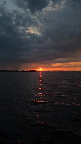 The video shows a sunset over water. The sky is filled with dark, ominous clouds, but a small sliver of orange sky is visible on the horizon where the sun is setting. The sun is reflected in the water, creating a bright line of orange light that stretches across the surface. The water is dark and choppy, with small waves breaking along the shore. The overall impression is one of beauty and tranquility mixed with a sense of impending storm.