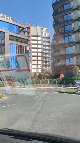 The video shows a street view with modern apartment buildings. There is a McDonald's restaurant visible between two tall buildings. A golden-colored car is parked on the side of the road. Overhead power lines and a large transmission tower are visible in the background against a clear blue sky. The street has yellow and black striped curbs and yellow painted road lines. Several small trees and bushes are planted along the sidewalk. A stop sign is visible. The footage appears to be taken from inside a vehicle.