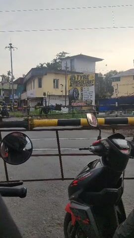 The video shows a view of a railroad crossing. A train is passing through the crossing, blocking traffic. The train cars are gray and red. People are visible inside one of the train cars. In the foreground, a motorcycle is parked behind a yellow and black barrier. A mirror attached to the motorcycle reflects the scene. A sign for "TYRE CONSULTANT" is visible on a building in the background. The sky is overcast.