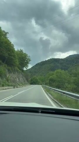 The video shows a view from inside a car driving on a rural road. The road has white lines marking lanes and a metal guardrail on the right side. On the left is a rock face covered with vegetation, and on the right, a mountain covered with trees. The sky is overcast with dark, grey clouds. The car is moving forward, as evidenced by the changing perspective. There is no visible interaction with other people or vehicles.