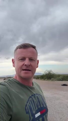 A man in a green Tommy Hilfiger t-shirt stands in a desert landscape under a cloudy sky. He appears to be recording a video of himself. The background shows a dirt road, sparse vegetation, and distant mountains. In some frames, vehicles like a white van and a blue truck are visible on a paved road to the left. The man smiles and speaks to the camera. The scene suggests an outdoor setting, possibly a roadside or a remote area.