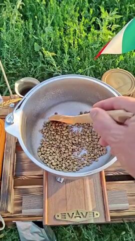 A close-up shot shows a hand holding a wooden spoon and stirring green coffee beans in a metal pot. The pot is placed on a wooden stand in an outdoor setting, with green grass visible in the background. An Italian flag is partially visible in the upper right corner of the frame. The coffee beans are light green and appear to be raw. The spoon is moving the beans around in a circular motion.