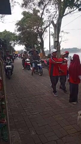 The video shows a paved walkway lined with trees on the right and what appear to be market stalls or small shops on the left. Several people are visible. In the foreground, a person on a black motorcycle with the license plate AE 4512 PL is riding towards the camera. Behind them, another person on a blue and white motorcycle is also moving forward. Further back, more motorcycles and people are present, including a red motorcycle with a rider. In the background, a body of water is visible, suggesting the location is near a lake or river. The atmosphere appears to be casual, possibly a street market or a recreational area.
