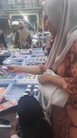The video shows a street scene with several people and food stalls. A woman in a pink hijab is at the forefront, looking towards a food stall. Multiple clear plastic containers filled with various colorful sweets and desserts are displayed on tables. There are other people, some wearing hijabs, browsing the food items. In the background, there are buildings and people on motorcycles, suggesting a busy urban or semi-urban environment. One person is handing something to another across one of the tables. The lighting appears to be overcast or late afternoon. The ground is paved with interlocking bricks.