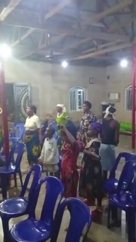 The video shows a group of people, including adults and children, in an indoor space. Some are standing and appearing to dance or move to music, while others are seated in blue plastic chairs. The room is lit with electric lights and has a simple interior, including a wooden ceiling structure. There are windows and a door visible, and a pole with red fabric wrapped around it stands near the center of the room. People are dressed in a variety of clothing styles, some in casual wear and others in more formal or traditional attire.