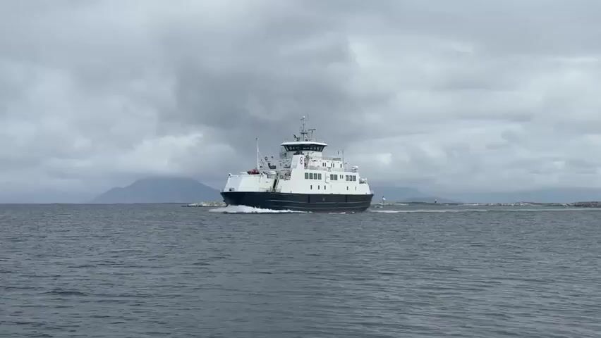 A wide shot shows a ferry traveling across choppy gray water under a cloudy sky. The ferry is white with a dark blue stripe around its hull, and it has a tall superstructure with windows. The ferry is moving from left to right in the frame and is creating a wake behind it. In the background, there are distant hills or mountains and a few small islands. The overall mood is somber and overcast.