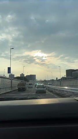 The video shows a view from inside a vehicle driving on a multi-lane highway. The time is likely late afternoon or early evening, as the sun is partially obscured by clouds, casting a diffused light. The sky is overcast with dramatic cloud formations. The highway is bordered by white sound barriers on both sides. In the distance, buildings and city structures are visible. The highway is moderately busy with traffic, consisting of cars and a truck. The camera is positioned behind the dashboard, looking forward through the windshield. The vehicle appears to be in the right lane.