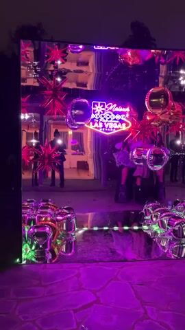 The scene is a nighttime outdoor event with a large mirrored wall as a focal point. The wall is decorated with pink starburst-shaped balloons, silver disco ball-shaped balloons, and a neon sign that reads "Maisun, Welcome to Fabulous Las Vegas, Nevada." The ground around the wall is covered with purple lighting, creating a vibrant, festive atmosphere. Several people are visible in the reflection of the mirror, including a person in all black taking photos, and others who appear to be posing or standing near the wall. The reflection in the mirror is slightly distorted, adding a surreal effect to the scene. The setting seems to be a garden or park, with trees visible in the background. The overall atmosphere is upbeat and celebratory, with music playing in the background.