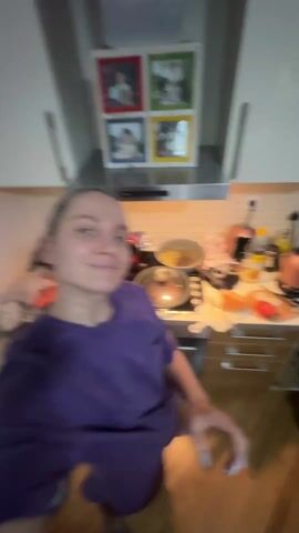 A woman in a purple t-shirt films herself in a kitchen. She has her hair in a ponytail and has a nose ring. There are several pots and pans on the stove behind her. There is a photo display above the stove. She looks surprised and puts her hands to her face. She then smiles.