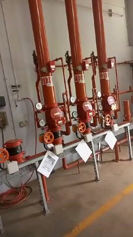 The video begins with a view of three orange fire suppression systems in a room with gray walls and a polished concrete floor. The camera pans to the left, revealing an orange toolbox and a white box on a metal stand, with a fan on the floor nearby. The camera then moves into a large, open warehouse space, following a group of people walking away from the camera. The group includes a person in a pink patterned dress, a person in an orange suit, and others in casual attire. There are stairs visible in the background, along with what appears to be a yellow forklift. The warehouse space is brightly lit with overhead fluorescent lights.