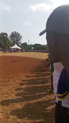 The video shows a crowd of people watching a race on a dirt field. The camera is positioned behind a group of onlookers, and the focus is on the runners in the distance. There is a white tent and a group of spectators near the finish line. The sky is partially cloudy.