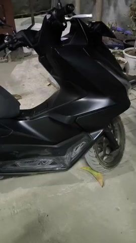 The video shows a close-up, side view of a black scooter parked indoors. The scooter is stationary. The floor is concrete. There are some wooden structures and a blue helmet visible in the background.
