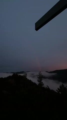 A time-lapse video shows a dusky sky over a misty mountain range. A faint rainbow is visible in the sky above the clouds. The top right of the frame is occupied by a dark, rectangular structure that appears to be part of a building or a cross. The trees and mountains are silhouetted against the dim light, and the clouds are thick and white, obscuring the lower slopes of the mountains.