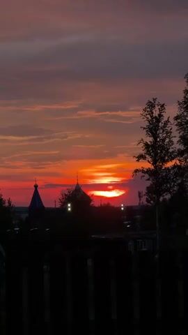 This is a vertical video clip featuring a sunset. The sky is a vibrant mix of orange and red hues, with streaks of darker clouds. In the foreground, silhouetted against the sunset, are trees and a wooden fence. In the middle ground, there are faint silhouettes of buildings, possibly including church domes, and some lights that suggest it is dusk. The overall impression is a peaceful, atmospheric sunset scene.