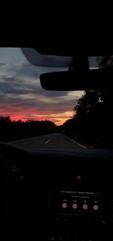 The video, shot from inside a car, shows the view through the windshield of a road at sunset. The sky displays shades of pink, purple and orange. Trees line both sides of the road, appearing dark in silhouette. The dashboard of the car is visible, including the steering wheel on the left, the hazard light button, and a digital display showing the text "Огр. скорости в стране/субьекте: Боснии и Герцеговине". The display also indicates speed limits of 50, 80, 100, and 130. The rearview mirror is prominent at the top center of the frame. The car is moving forward on the road.