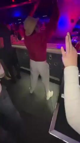 The video takes place inside a crowded nightclub. A man wearing a red jacket, light pants, a beige hat, and white sneakers is seen dancing in front of a bar. He waves a large white balloon or cloth in the air.  Other people stand nearby, some with their hands raised in the air. The club is lit with red and blue lights and a disco ball hangs from the ceiling. Some people are standing and others are moving through the crowd.