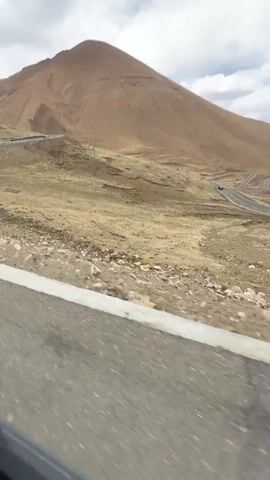 The video shows a landscape of hills and mountains under a cloudy sky. A winding road stretches into the distance, and a black car is visible on the road. The foreground consists of rocky and dry terrain beside the road.