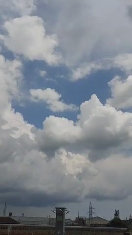 The video displays a time-lapse of the sky from Romanivka, Ukraine. The scene is dominated by a dynamic sky filled with a mix of fluffy white cumulus clouds and darker, more ominous storm clouds. Patches of bright blue sky peek through, suggesting a changing weather pattern.

In the foreground, the rooftops of industrial or commercial buildings are visible, along with a tall transmission tower and other structures that indicate an urban or semi-industrial environment. A lone, well-maintained tree with lush green foliage is positioned on the right side of the frame, its leaves rustling gently, contrasting with the vastness of the sky. The camera seems to be mounted at a height, possibly from a building or elevated platform, offering a panoramic view.

The time of day appears to be daytime, likely late morning or afternoon, given the brightness of the sky and the shadows cast by the clouds. The weather is unsettled, with the presence of both bright clouds and darker storm clouds, suggesting a period of transition. The overall mood is one of natural grandeur and the ever-changing moods of the atmosphere. There is no visible text in the media file.