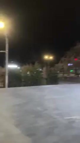 The video shows a nighttime street scene, possibly in a city. The camera slowly pans across a gray stone walkway toward a modern building with large glass windows. Behind the building, there are trees and other buildings, illuminated by streetlights. A white van is parked near a traffic light in the background. The overall impression is one of urban tranquility at night.