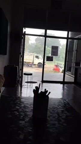 The video opens with a dimly lit interior of what appears to be a business or shop, looking out through large glass doors. The flooring is dark and reflective, with a patterned design. In the foreground, a silhouette of a cylindrical object holding several items, possibly pens or pencils, is visible. To the left, a dark cylindrical object, perhaps a trash can or a large container, is partially visible. Just outside the glass doors, a light-colored truck with a beige cargo bed is parked. Beside the truck, a motorcycle is parked. Through the doors, it's evident that it is raining, with raindrops visible on the glass and a generally wet environment outside. Green trees and vegetation form the backdrop. A round, metal stool is positioned near the doors. There are signs posted on the glass doors. The overall impression is a view from inside a building during a rainy day, looking out at a parked truck and motorcycle.